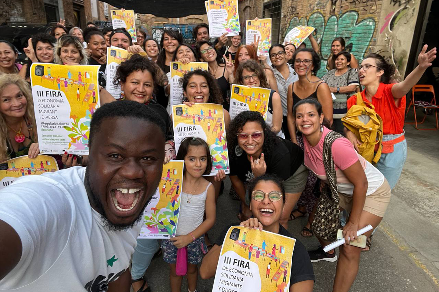Participantes de la Fira d'Economia Solidària Migrant i Diversa con los carteles del evento en Barcelona